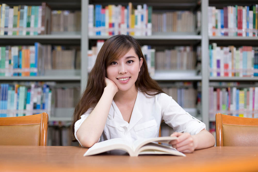 Beautiful asian female student read book in library with bookshelf as background.